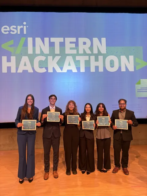 Krishna Panjiyar, far right, on stage with five fellow Esri interns, each holding a certificate in front of the Esri Intern Hackathon banner